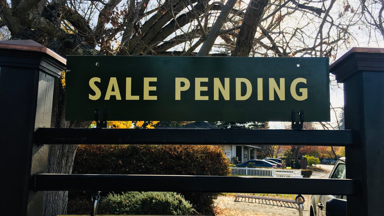 A green and yellow Sale Pending sign on a fancy post with trees, bushes, and a white picket fence in the background.; Shutterstock ID 1876850083