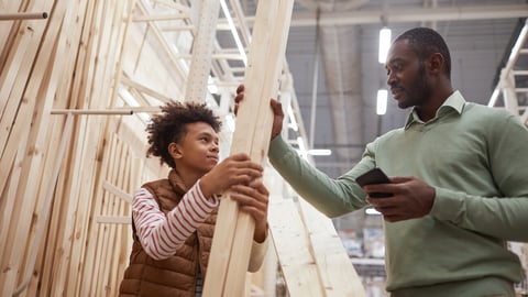 Low angle portrait of African-American father and son shopping together in hardware store, focus on man choosing wooden boards for construction or home improvement; Shutterstock ID 1891257115