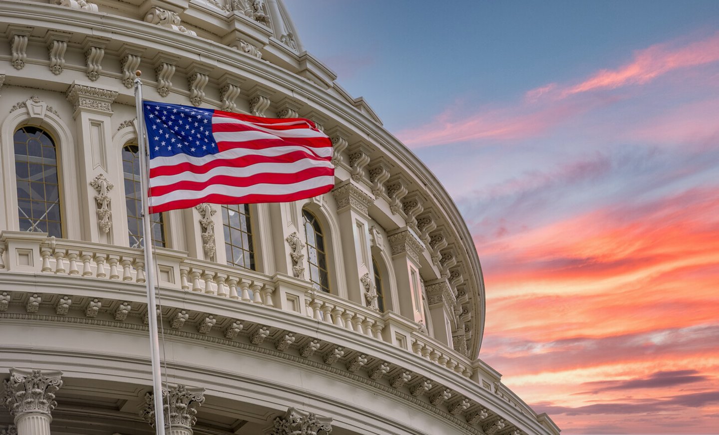 View of the United States Capitol Rotunda Dome in Washington DC with the Star Spangled American Flag against colorful dramatic sunset sky background; Shutterstock ID 1814961938