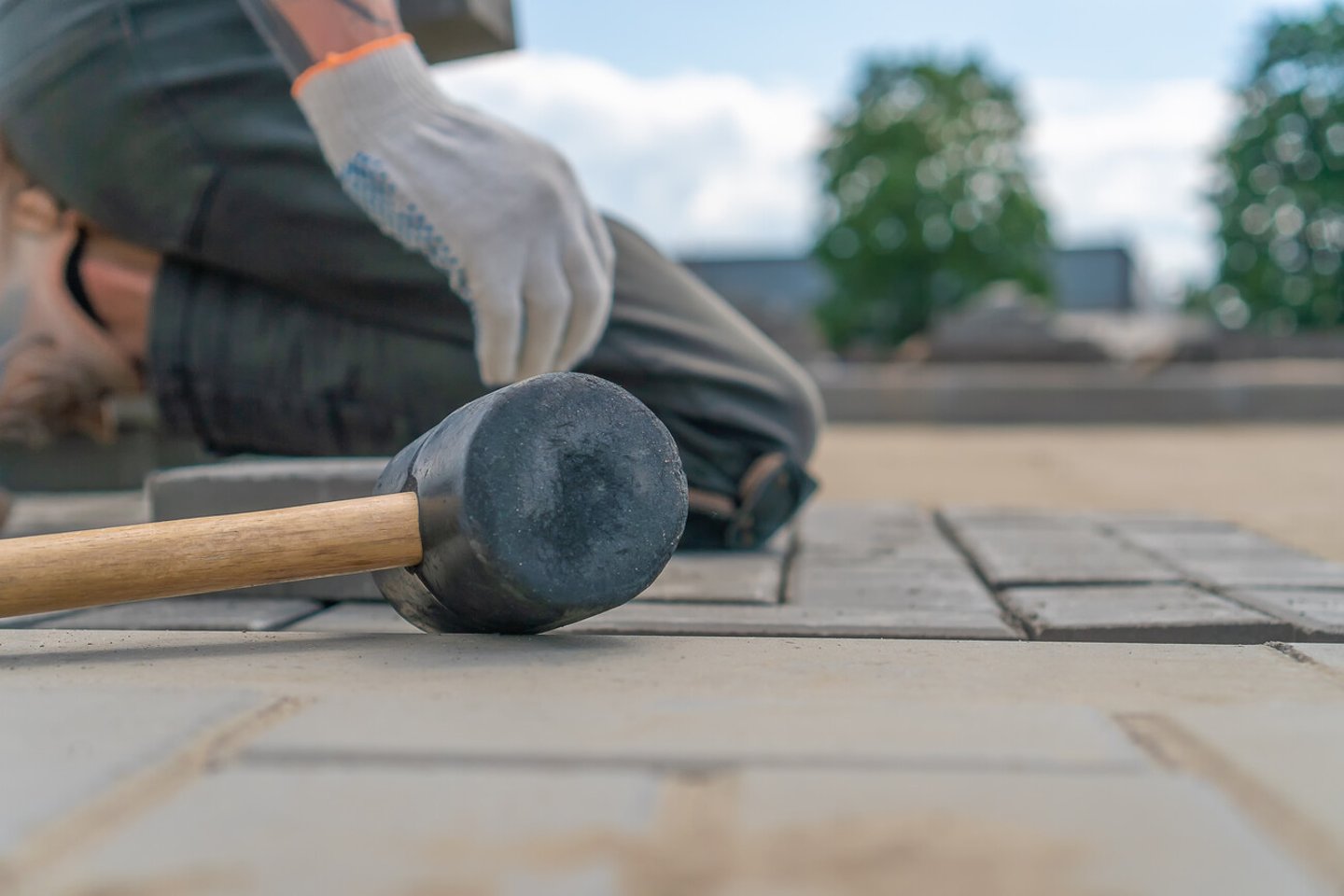 Close-up of a construction worker's gloved hands laying outdoor paving slabs on a prepared foundation on a summer sunny day, focus on a construction paving hammer; Shutterstock ID 2000459915